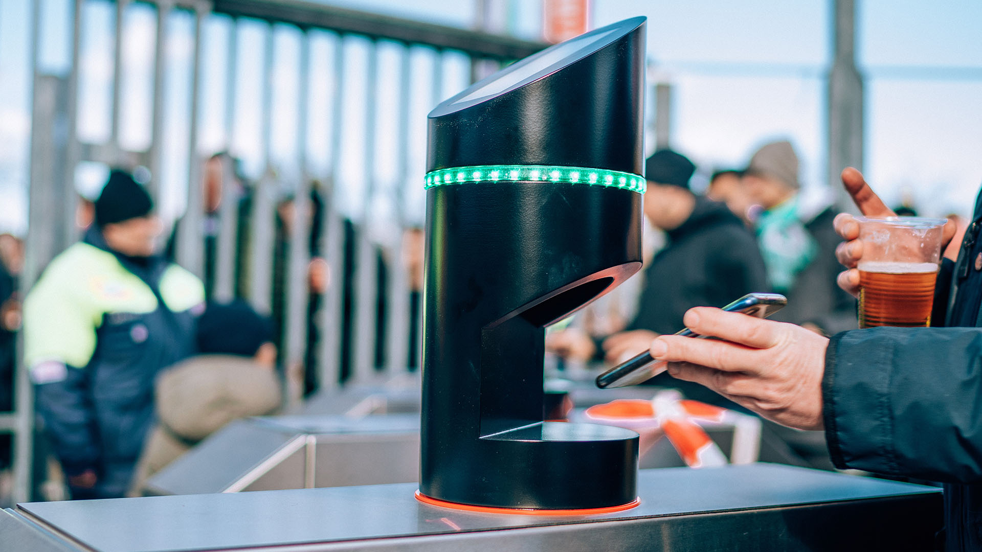 Die Hand einer nicht erkennbaren Person hält ein Handy unter einen Ticketscanner, im Hintergrund ein Zaun und weitere Menschen.