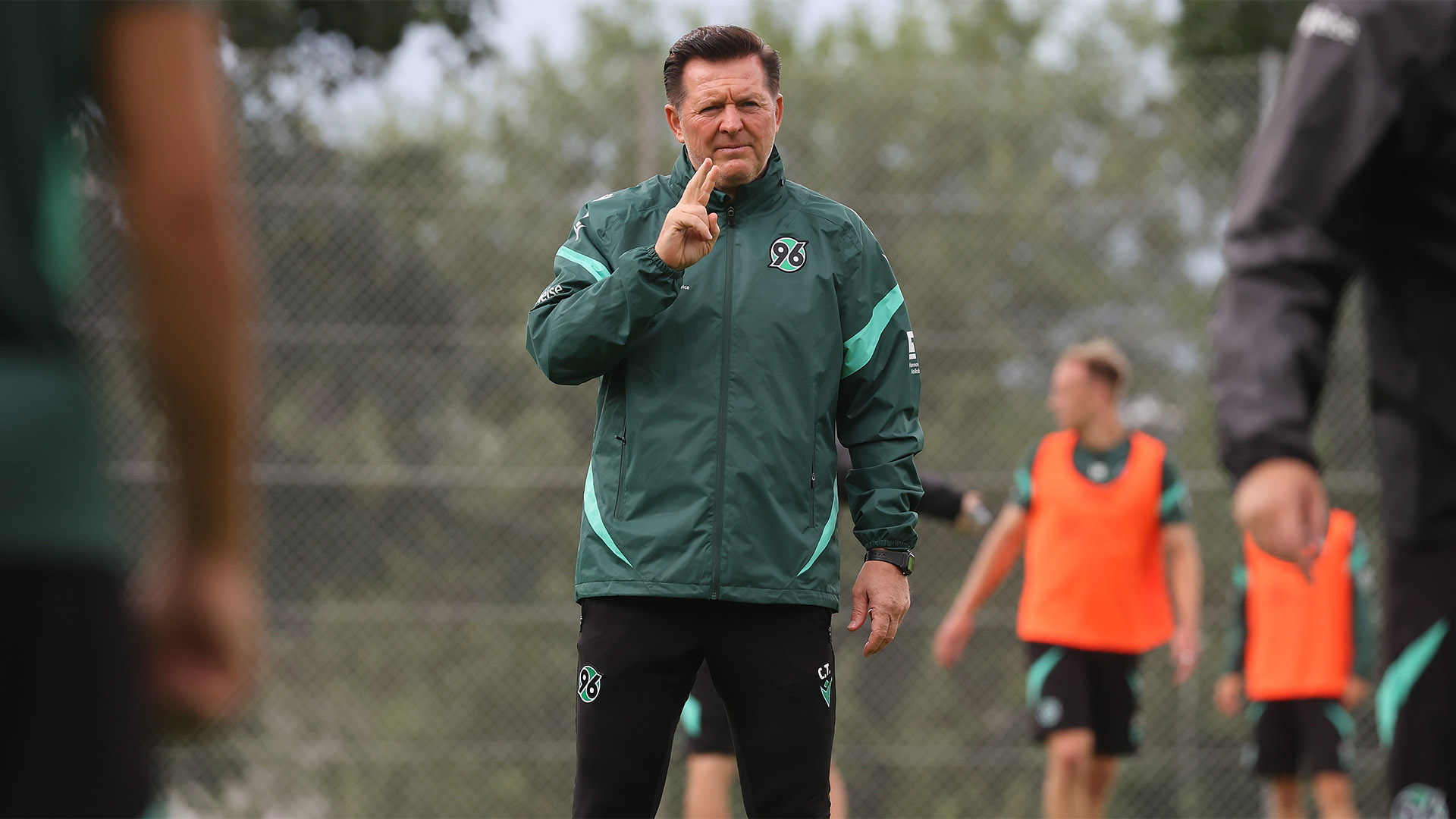 Christian Titz steht in grüner Regenjacke auf dem Trainingsplatz und zeigt mit dem Zeige- und Ringfinger der rechten Hand.