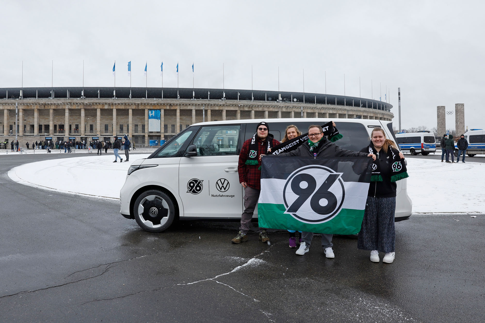 Zwei Frauen und zwei Männer stehen vor dem weißen ID. Buzz und halten eine 96-Fahne, im Hintergrund das Olympiastadion.