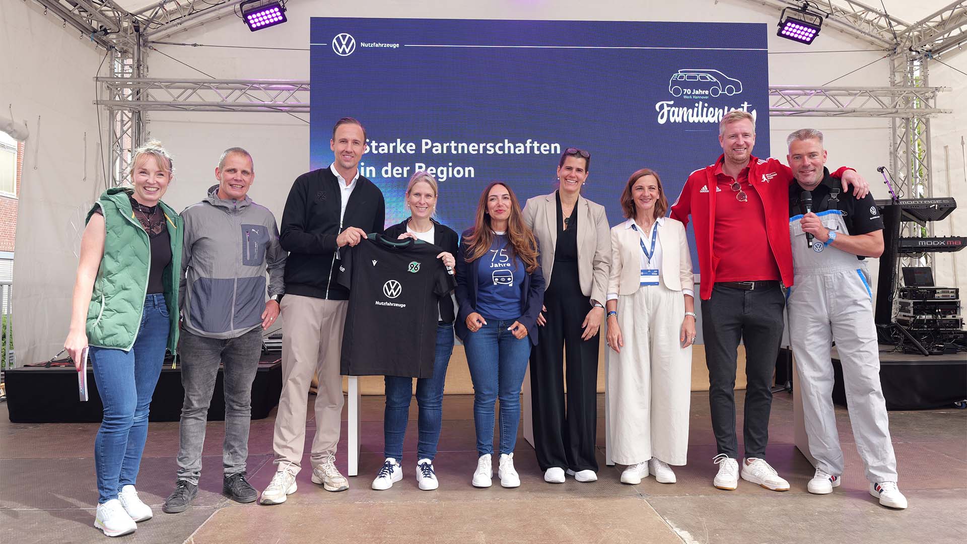 Neun Männer und Frauen stehen auf einer Bühne und halten ein schwarzes T-Shirt mit dem VW-Logo und dem 96-Logo hoch.