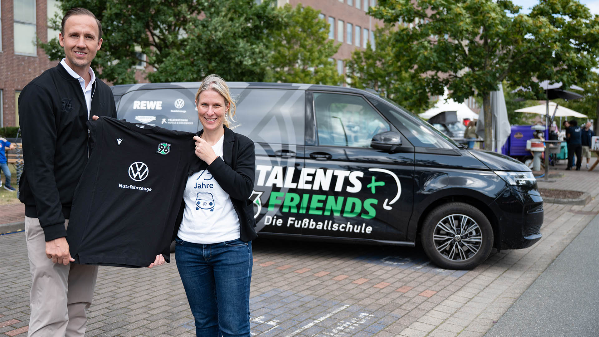Ein Mann und eine Frau stehen vor einem Bulli und halten ein schwarzes T-Shirt mit dem VW-Logo und dem 96-Logo hoch.