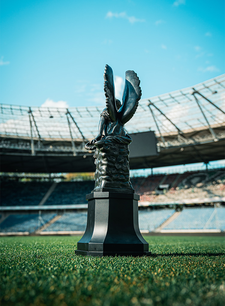 Die Trophäe, eine Frau mit Flügeln, ist von hinten zu sehen, im Hintergrund das Spielfeld der Heinz von Heiden Arena sowie die Ränge im Norden. 