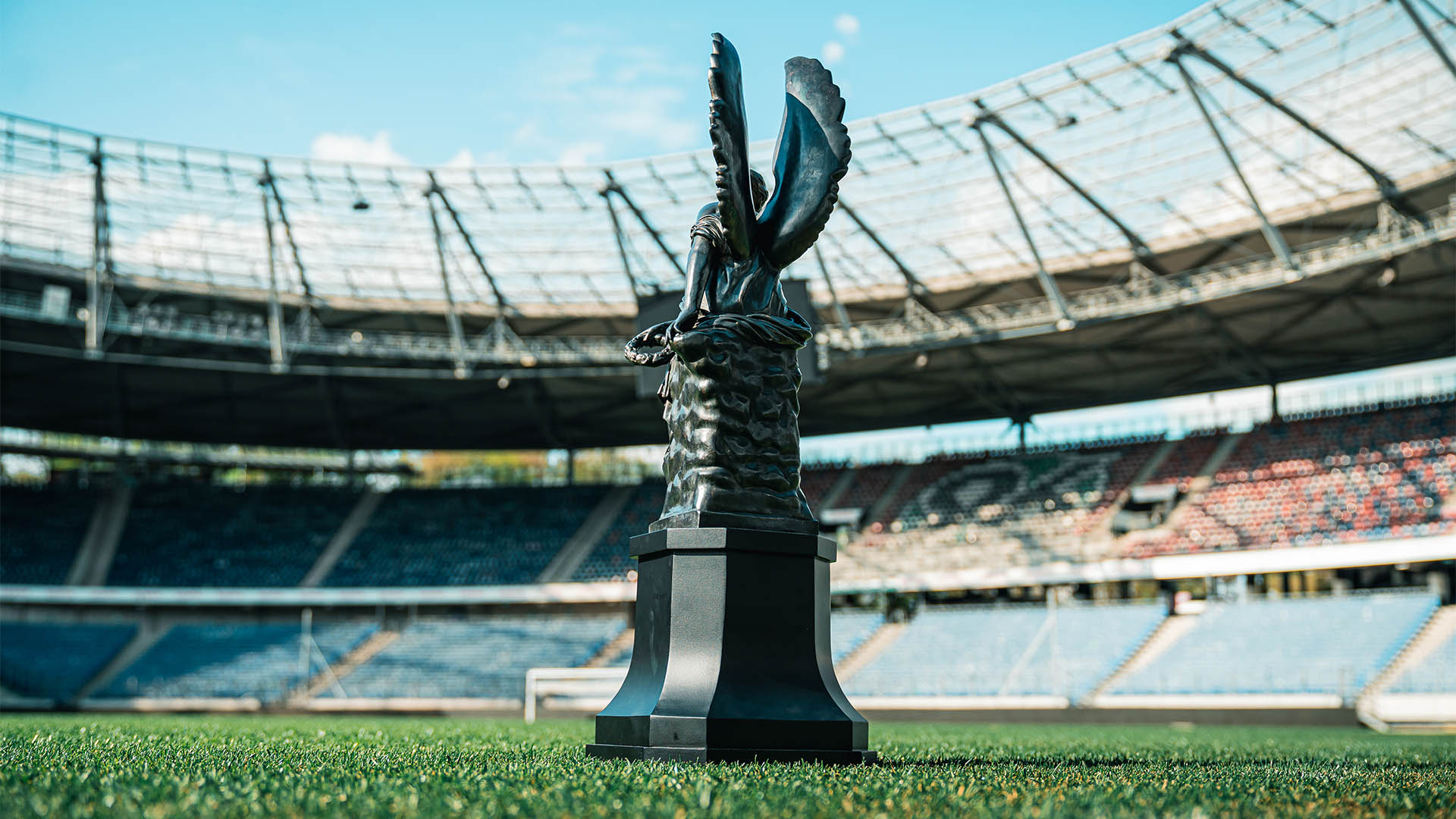 Die Trophäe, eine Frau mit Flügeln, ist von hinten zu sehen, im Hintergrund das Spielfeld der Heinz von Heiden Arena sowie die Ränge im Norden. 