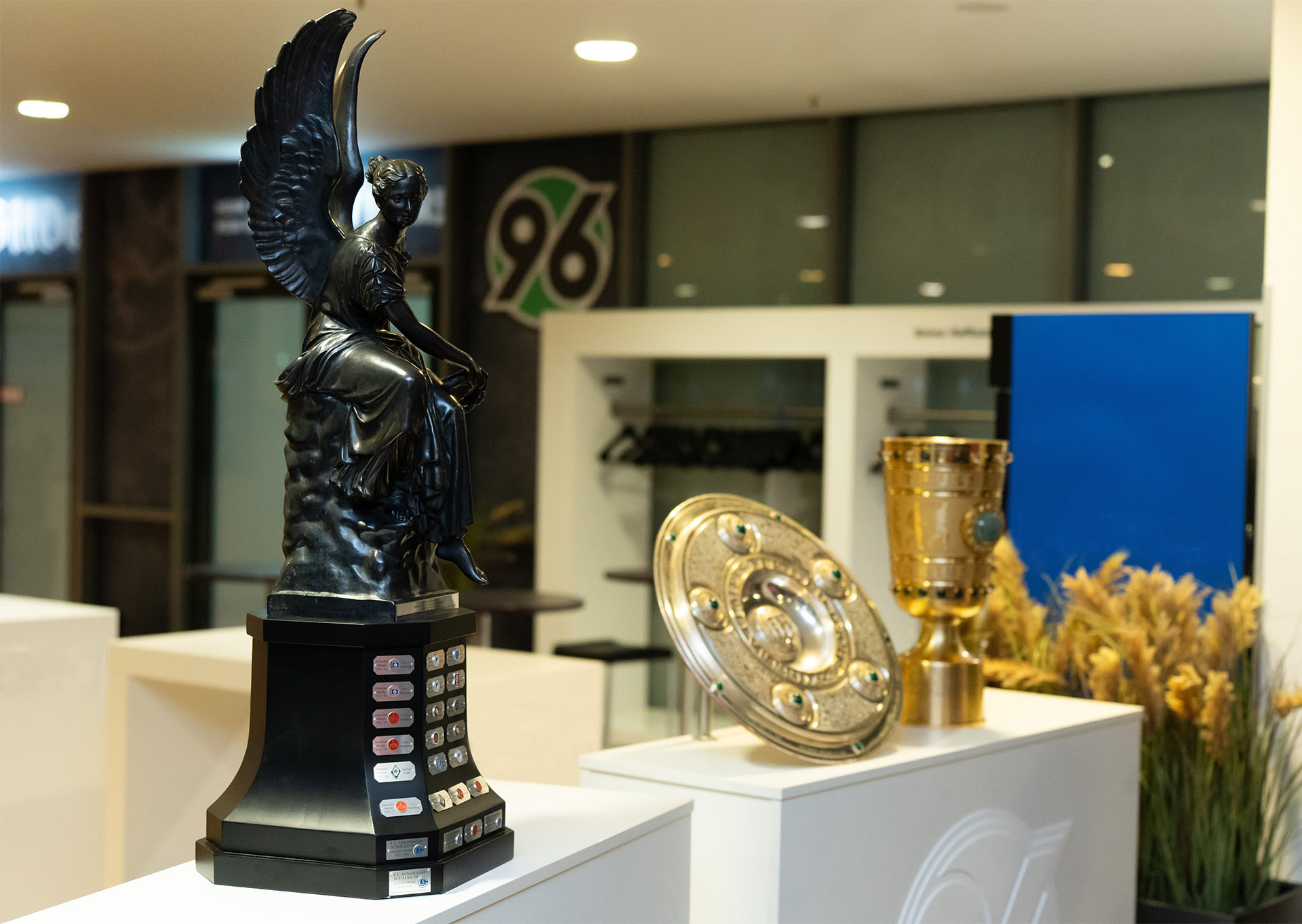 Die Viktoria steht neben der Meisterschale und dem Pokal auf einem Podest.
