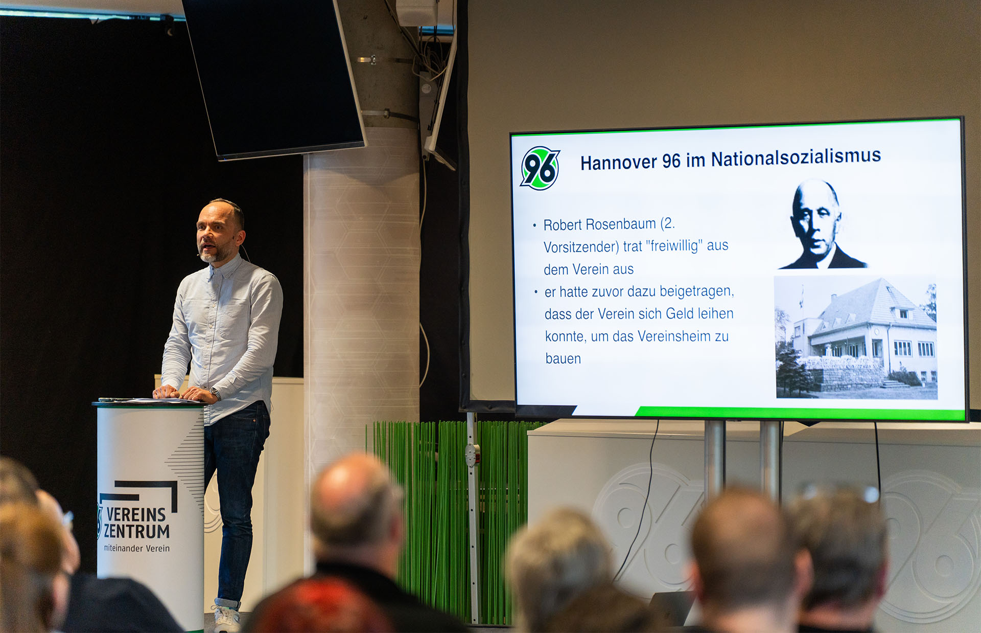 Blick auf die Bühne, dort steht ein Mann an einem Pult und spricht, neben ihm ist eine Präsentation mit dem Titel "Hannover 96 im Nationalsozialismus" auf einem großen Bildschirm zu sehen
