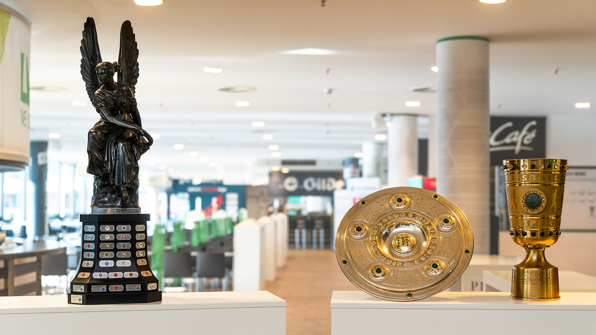 Die Viktoria steht neben der Meisterschale und dem Pokal auf einem Podest.