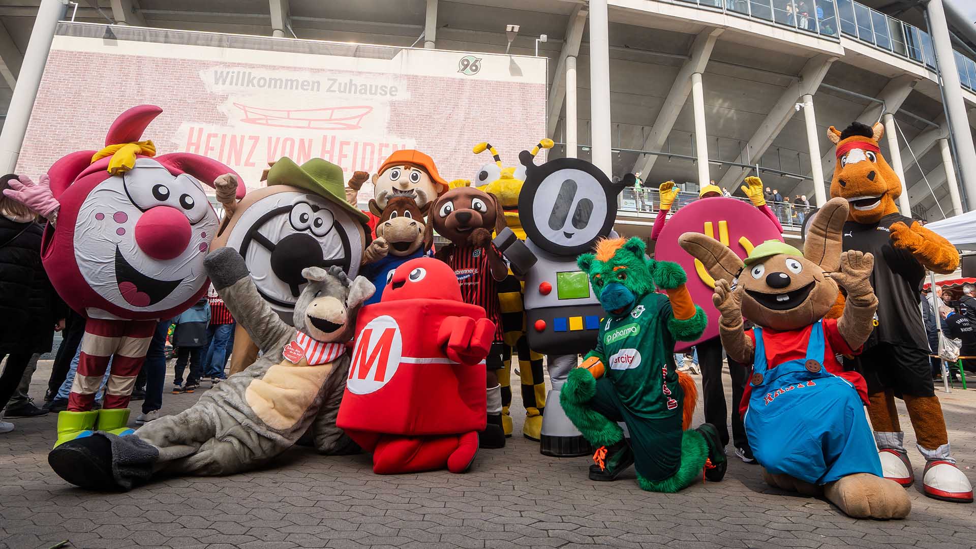 13 Maskottchen die vor einem Stadion stehen.