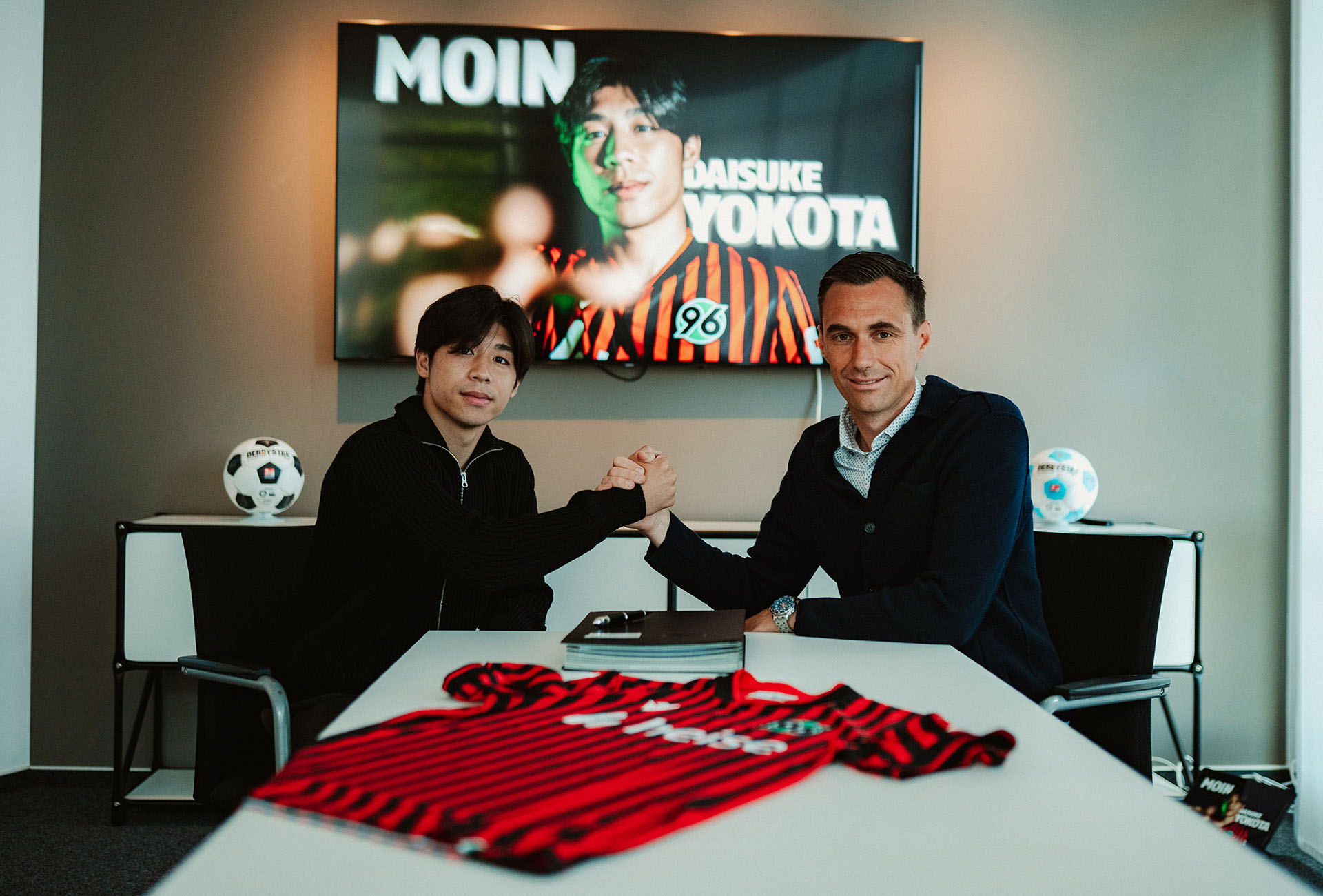 Daisuke Yotoka unterschreibt am Schreibtisch von Marcus Mann, Mann sitzt daneben, beide lächeln.