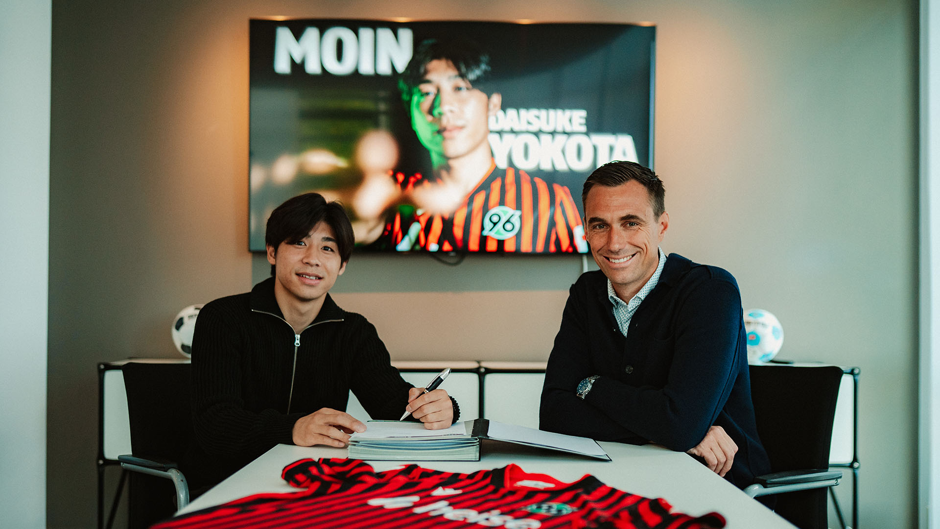 Daisuke Yotoka unterschreibt am Schreibtisch von Marcus Mann, Mann sitzt daneben, beide lächeln.