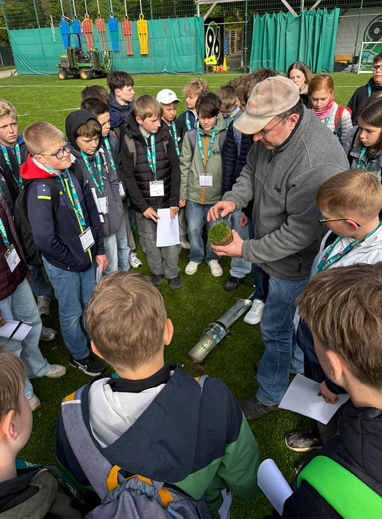 Die Kinder stehen im Kreis um einen Mann, der ein Stück Rasen in der Hand hält. (Foto: 96/Redaktion)