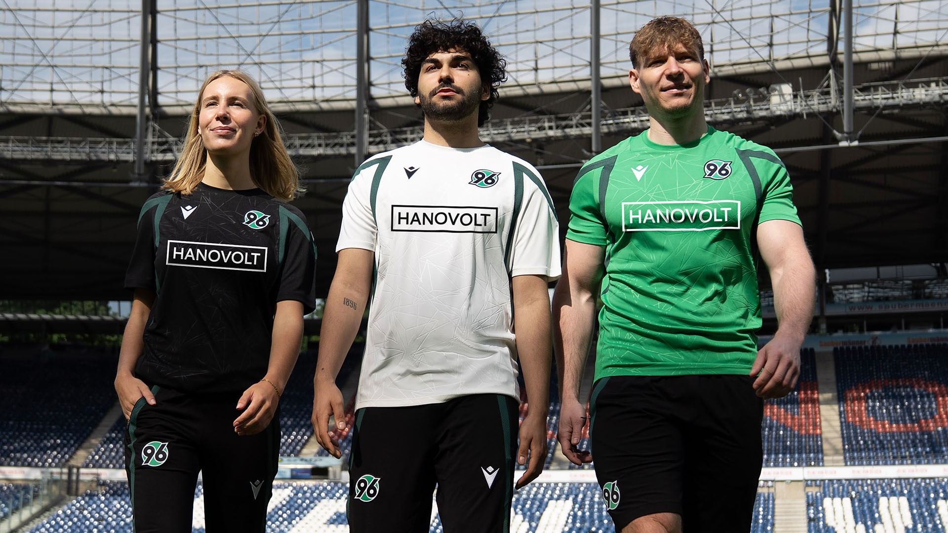 Eine Frau und zwei Männer in schwarzer, weißer und grüner Trainingskleidung im Stadion.