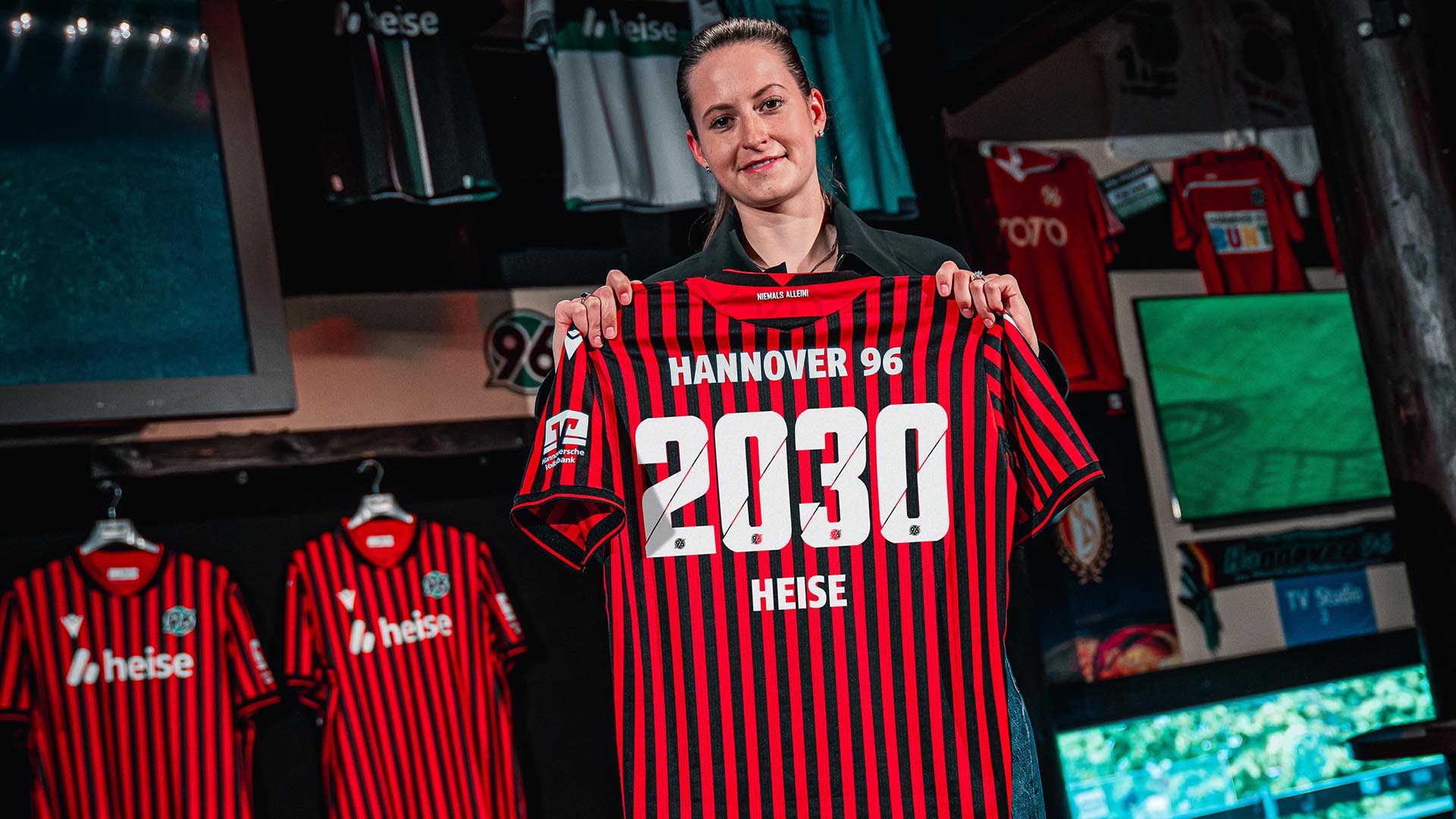 Eine Frau mit zusammengebundenen Haaren hält ein 96-Heimtrikot mit dem Flock "Hannover 96, 2030, heise".