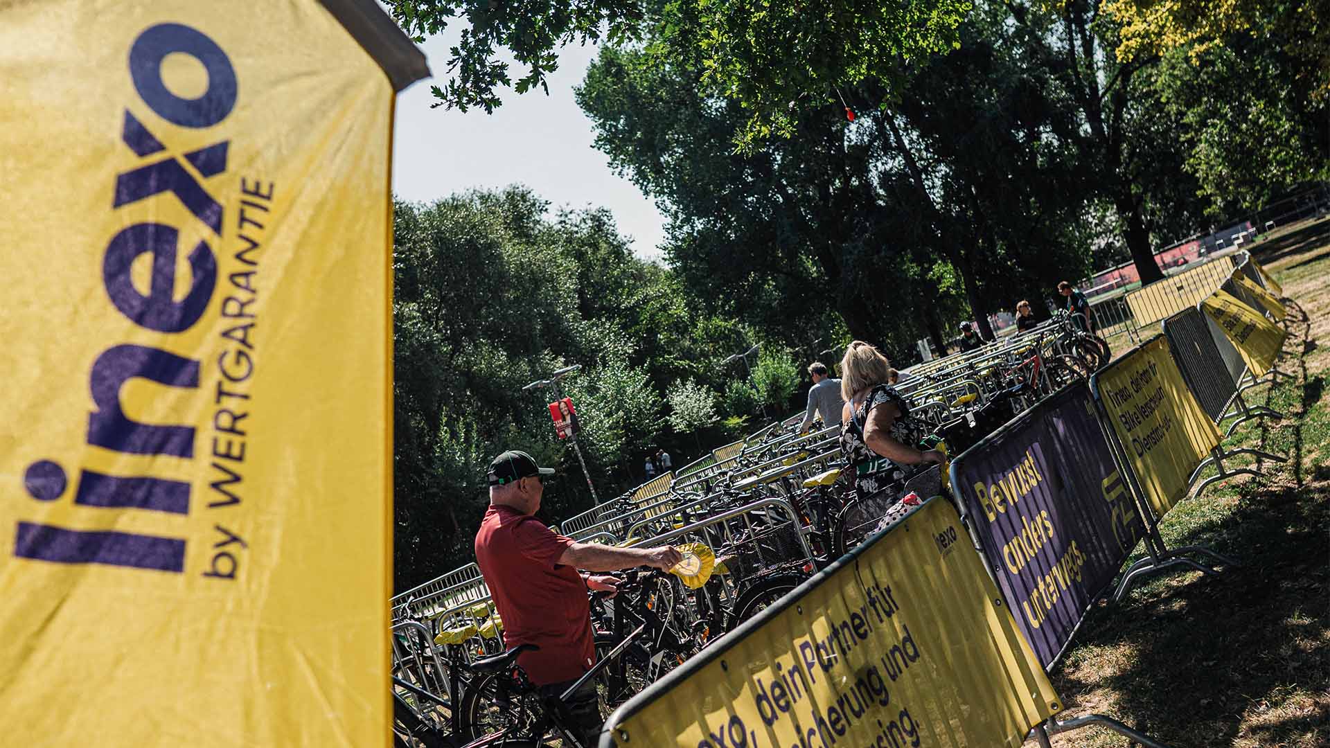 Eine linexo-Beachflag steht im Vordergrund, dahinter ist ein Fahrradstellplatz zu sehen, auf dem zwei Menschen gerade ihr Rad parken.