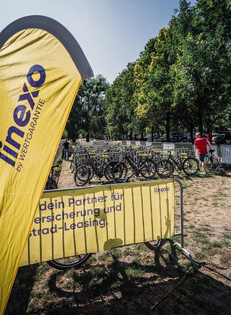 Blick auf einen Fahrradstellplatz auf einer Wiese, im Vordergrund steht eine gelbe linexo-Beachflag.