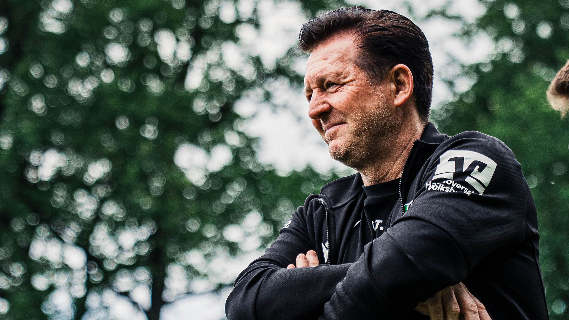 Christian Titz mit verschränkten Armen und einem Lächeln am Spielfeldrand.