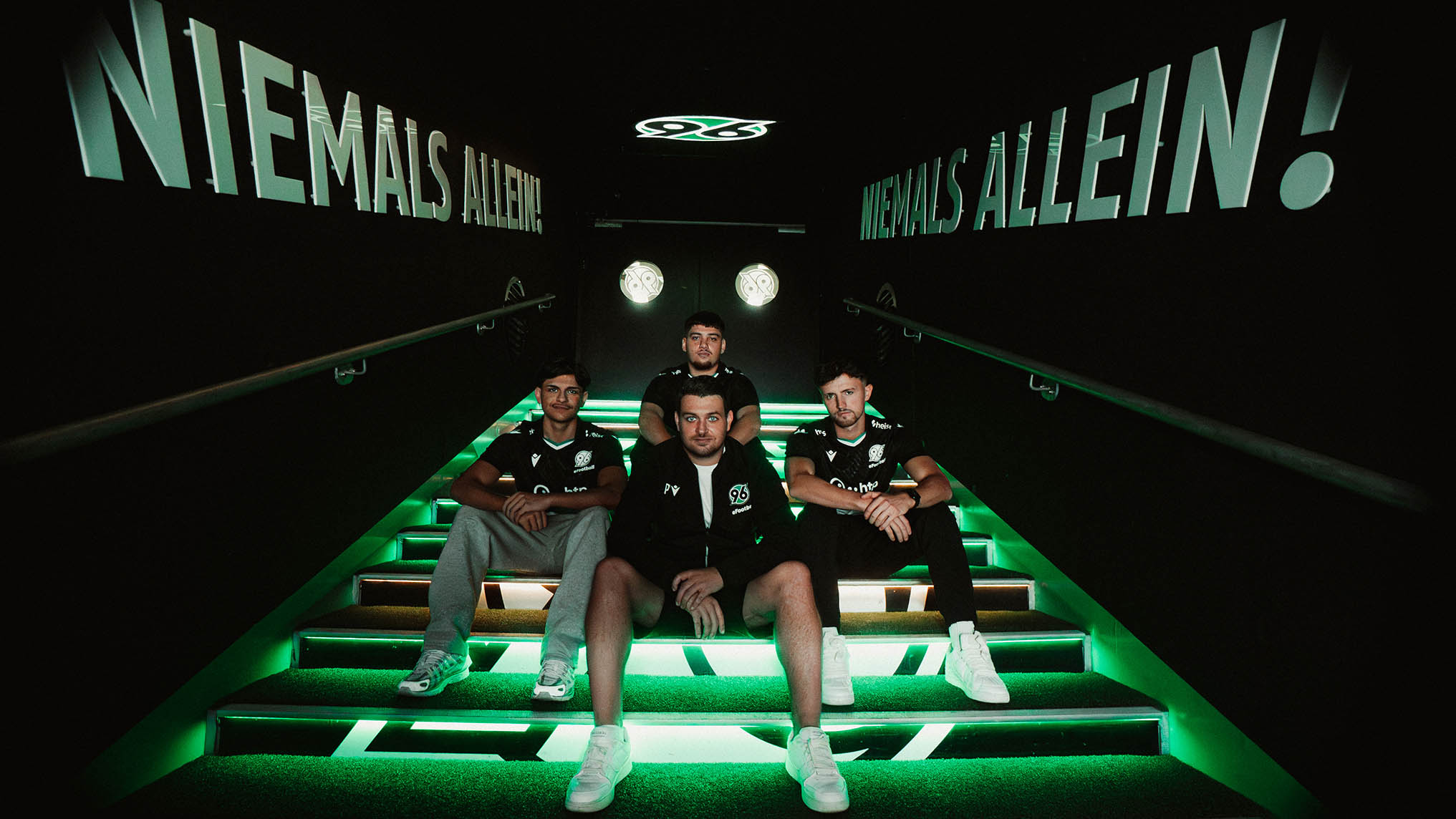 Drei 96 E-Footballer und deren Coach sitzen auf Treppen des Spielertunnels.