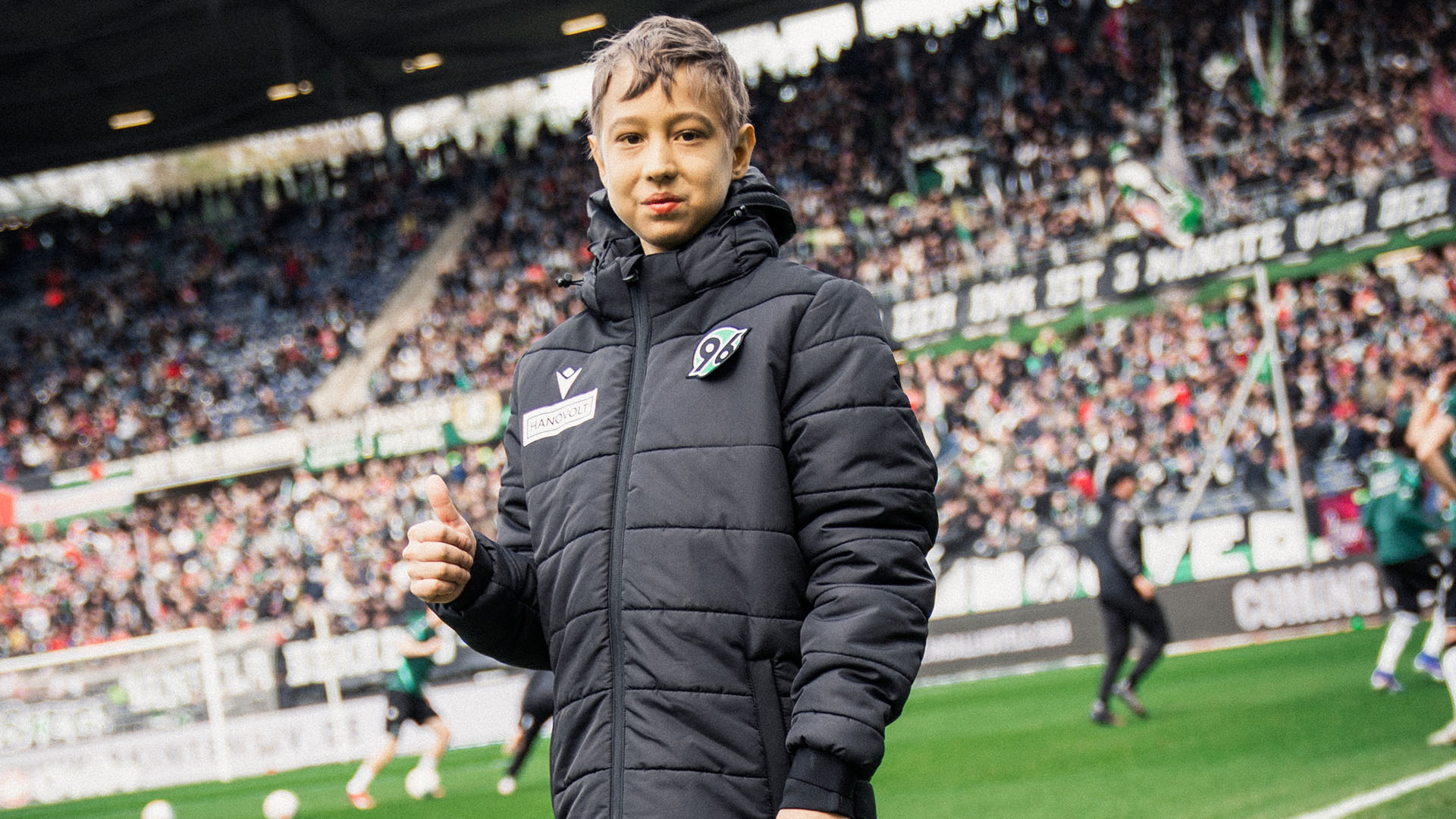 Vladislav steht in einer schwarzen Jacke am Spielfeldrand mit erhobenem Daumen.