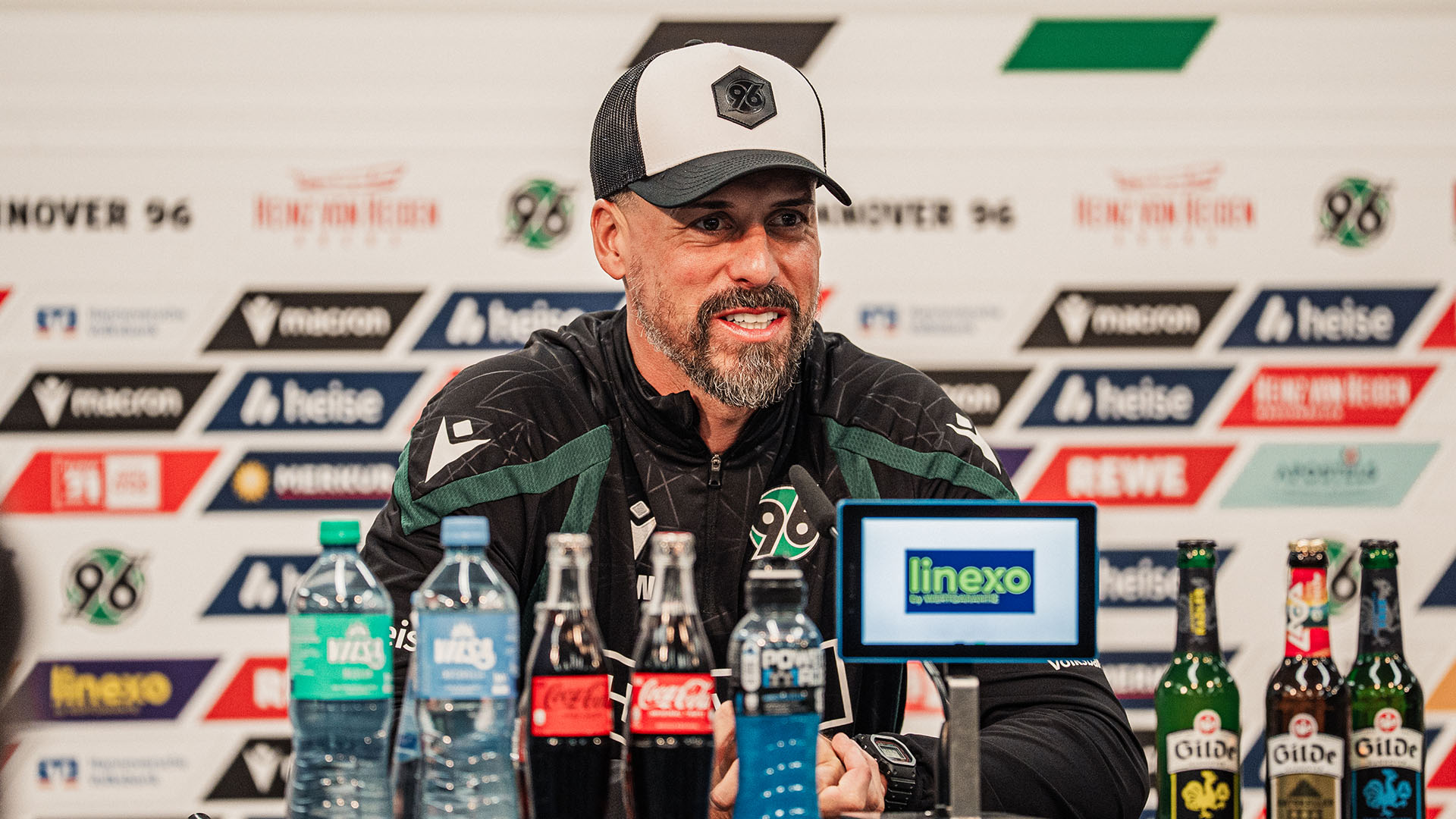 Der 96-Cheftrainer Sandro Wagner sitzt auf dem Pressekonferenz-Podium, hinter ihm unscharf eine Sponsorenwand.